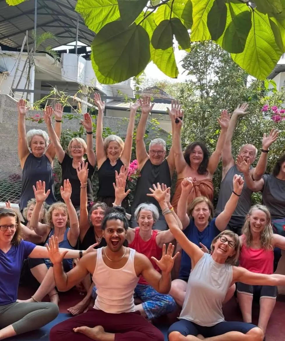 Nithin sits cross-legged on a mat, laughing, in front of a big group of students raising their arms under a canopy of green leaves