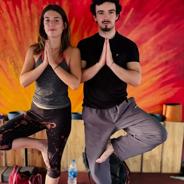 A woman and a man balance in tree pose, palms together, in front of the painted sun