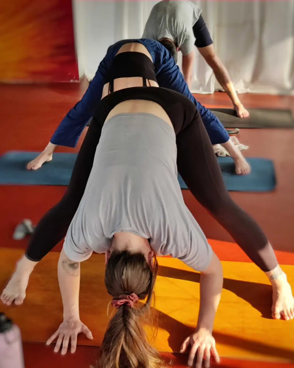 A student folds forward into a wide-legged stance on an orange mat in morning sunlight